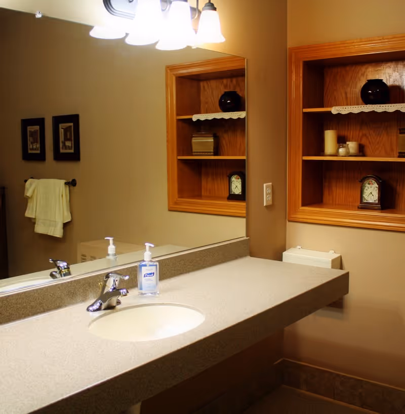 Bathroom sink area with a large mirror above a beige countertop. A chrome faucet and a bottle of hand sanitizer are on the countertop. On the wall to the right, there is a wooden recessed shelf with decorative items including a clock, candles, and a black vase. A towel rack with a white towel and two framed pictures are visible on the left wall.