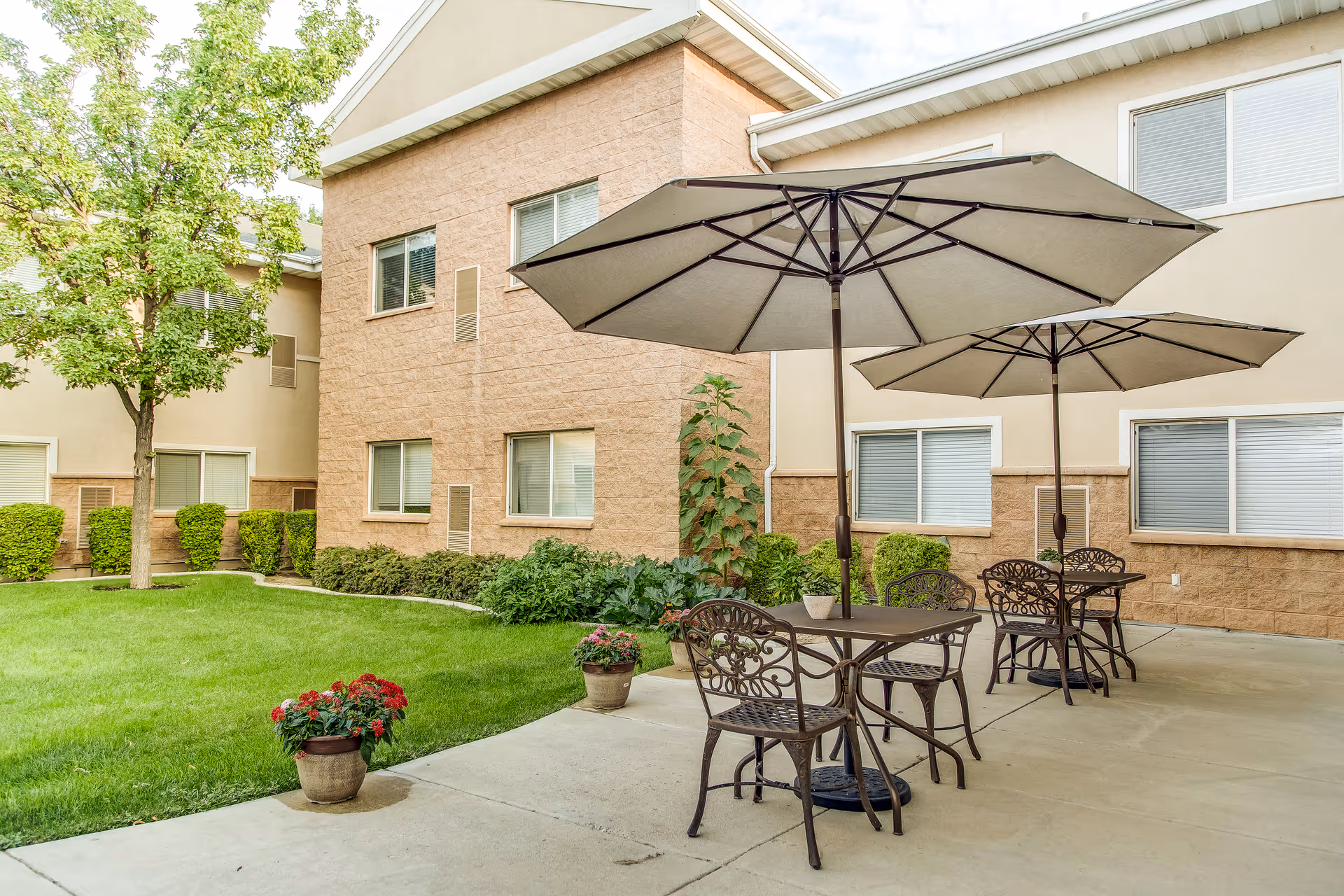 Outdoor patio area with metal tables and chairs under large beige umbrellas, surrounded by a green lawn, potted plants, and a two-story building with windows in the background.