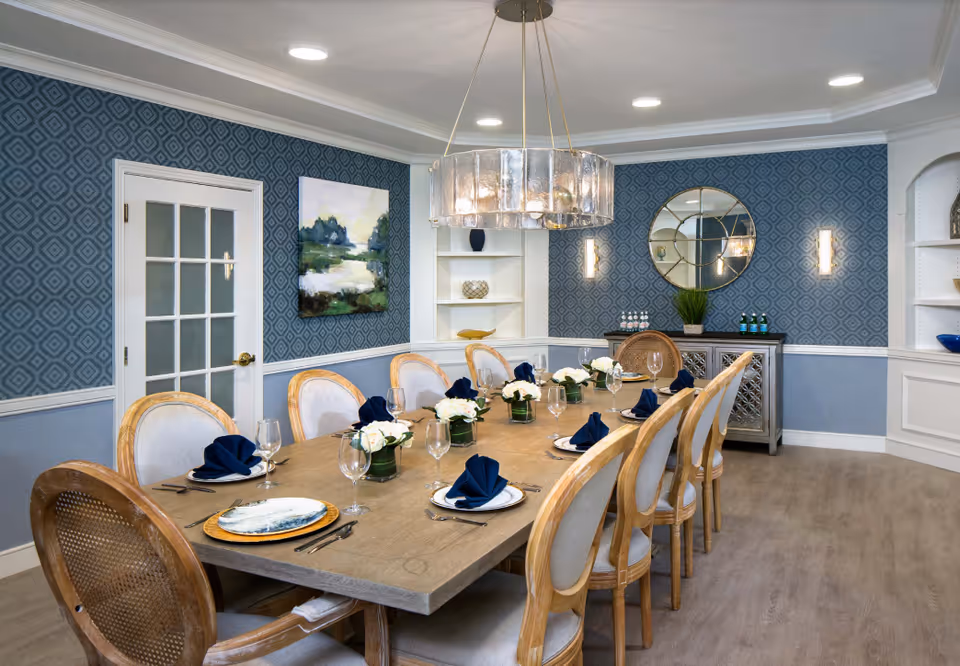 A well-decorated dining room with a long wooden table set for eight people. The table has white plates with blue napkins, wine glasses, and small flower arrangements. The room features blue patterned wallpaper, a large round mirror on the wall, a modern chandelier hanging above the table, and built-in white shelves with decorative items.