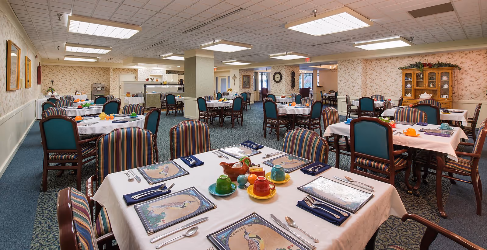 A spacious dining room with multiple tables covered in white tablecloths, each set with colorful cups, saucers, placemats, and silverware. The chairs have striped upholstery and wooden frames. The room has floral wallpaper, carpeted floors, and bright overhead lighting. A wooden cabinet with decorative items is visible against the far wall.