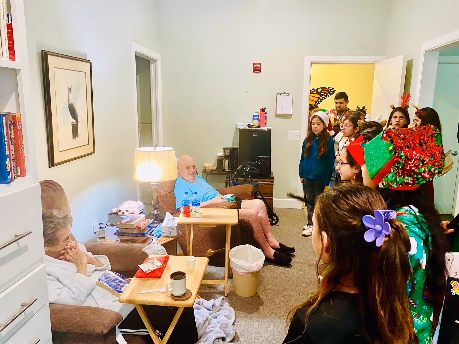 A group of children dressed in festive holiday attire, including Santa hats and reindeer antlers, standing in a room with two elderly people seated in armchairs. The elderly man is sitting with a walker nearby, and the elderly woman is seated with a blanket. The room has a lamp, books, and a framed picture on the wall.