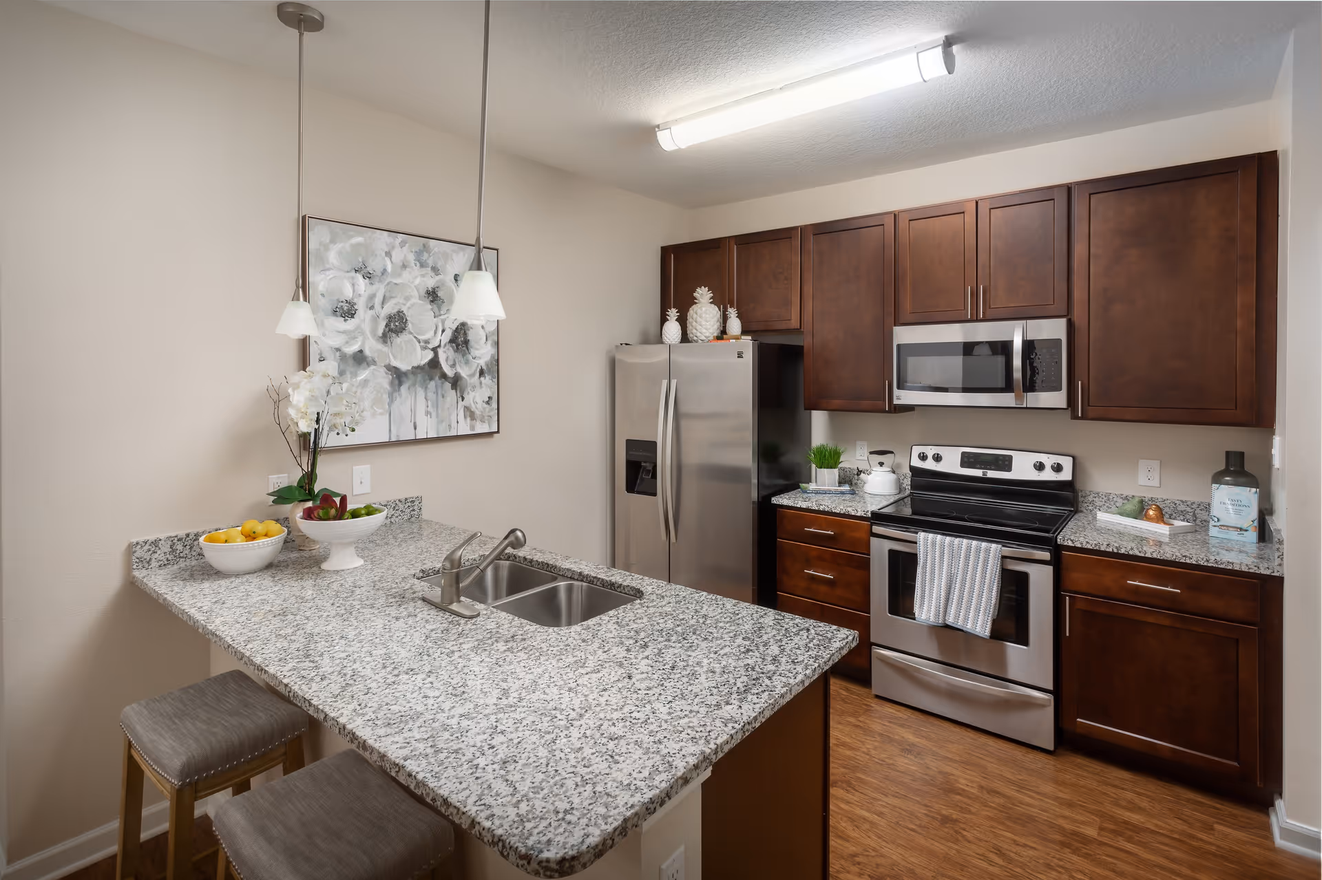 Modern kitchen with granite countertops, dark wooden cabinets, stainless steel refrigerator, stove, and microwave. Two pendant lights hang over a breakfast bar with two cushioned stools. Decorative items include bowls of fruit, a potted plant, and a floral painting on the wall.