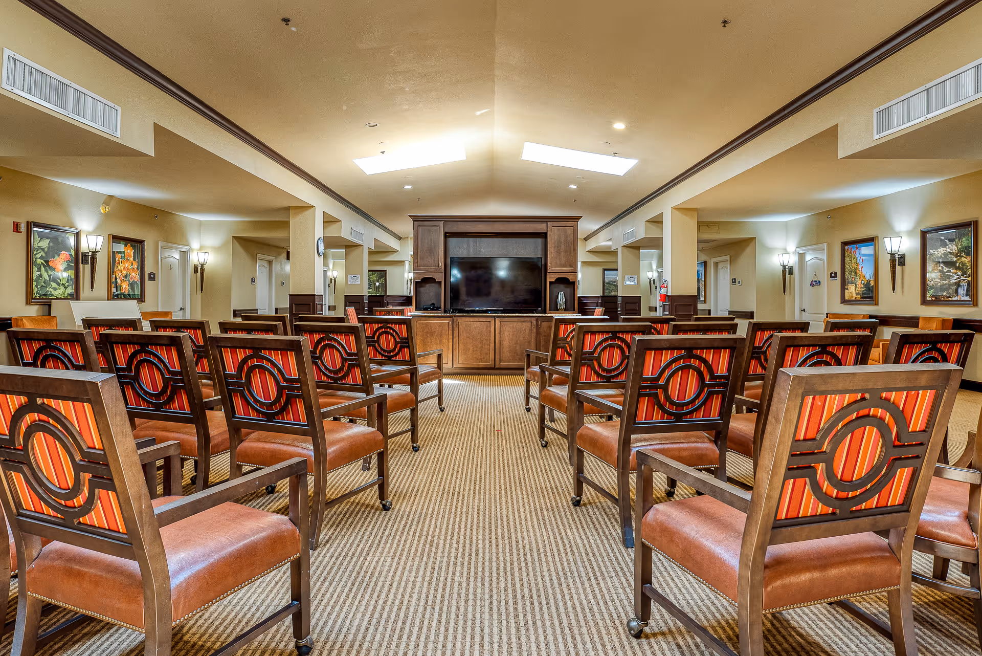 A spacious common area with rows of wooden chairs featuring red and orange striped cushions, facing a large flat-screen TV mounted on a wooden cabinet. The room has beige walls, carpeted floor, wall sconces, framed artwork, and skylights on the ceiling providing natural light.
