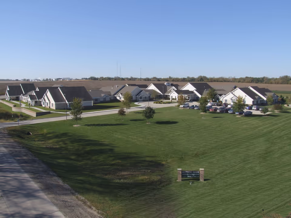Aerial view of Kentucky Ridge senior living neighborhood showing multiple single-story buildings with pitched roofs, a parking lot with cars, well-maintained green lawns, and a clear blue sky.