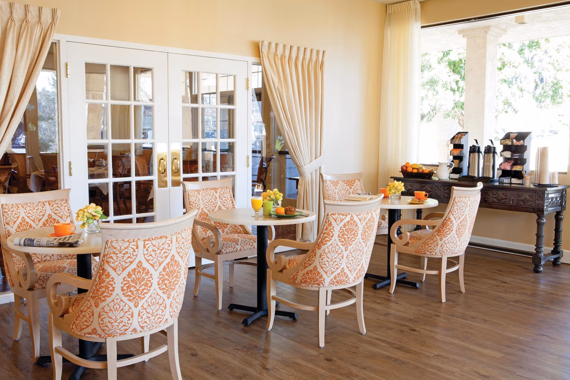 Sunny dining room with round tables, patterned upholstered chairs, and a beverage station by large windows.