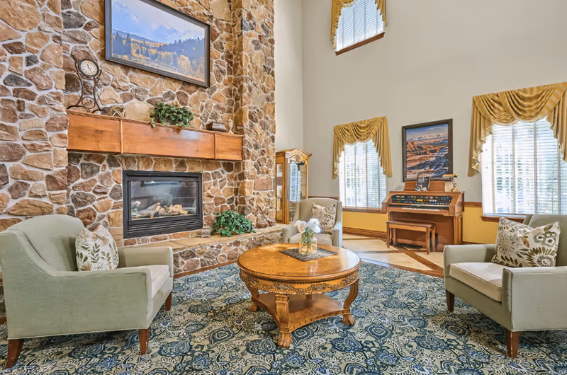 A cozy living room with a stone fireplace, armchairs arranged around a round wooden coffee table, and tall windows with gold drapes.