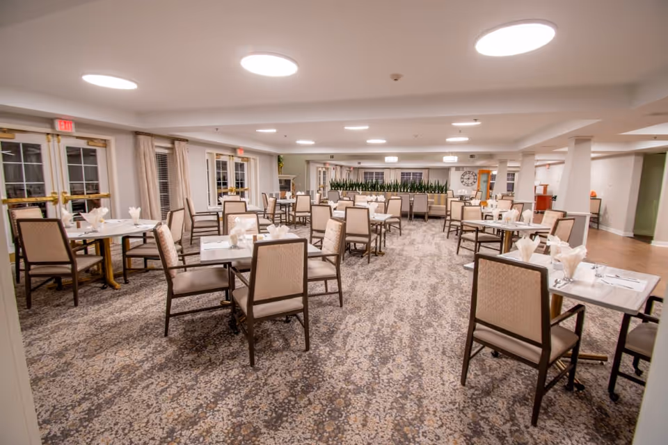 A spacious dining room with multiple tables and chairs arranged neatly. Each table is set with napkins and cutlery. The room has patterned carpet flooring, large windows with curtains, and bright ceiling lights. There are plants along the far wall and a large clock on the right wall.