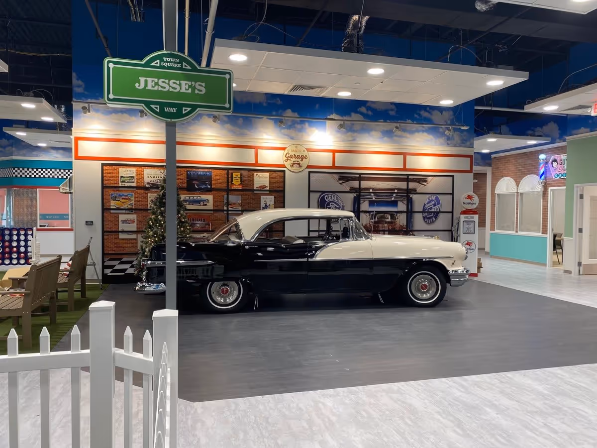 Indoor scene of a senior living facility area named Jesse's Way at Town Square University Parkway, featuring a vintage black and white car displayed on a gray floor. The background includes a wall decorated with vintage automotive signs and a Christmas tree. There are benches and a large Connect Four game to the left, and a barber shop area to the right with a neon barber pole sign.