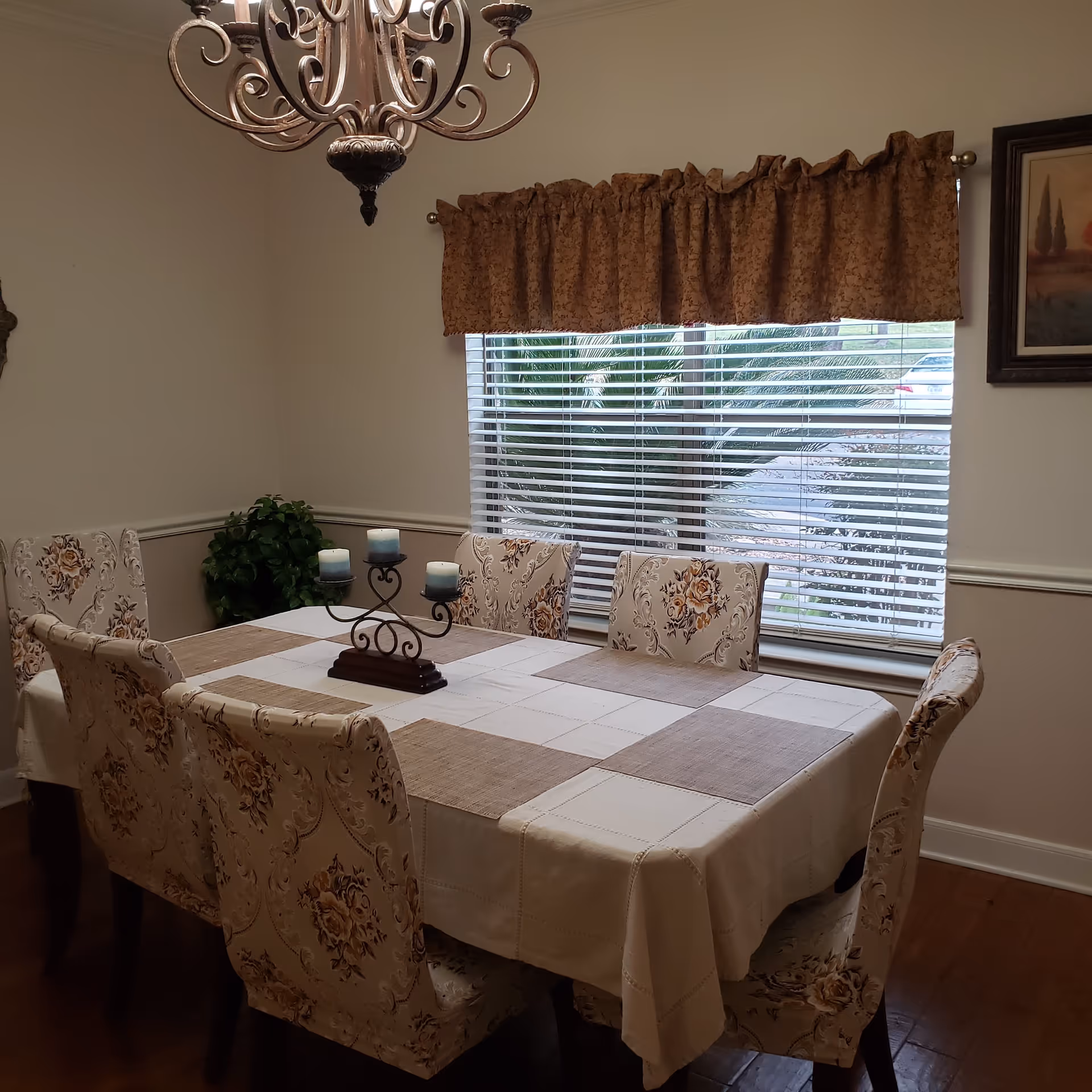 A dining room with a rectangular table covered by a beige and brown checkered tablecloth. Six upholstered chairs with floral patterns surround the table. A decorative candle holder with four blue candles is placed in the center of the table. A window with white blinds and a brown valance is behind the table, and a chandelier hangs above. There is a green plant in the corner and a framed painting on the wall.