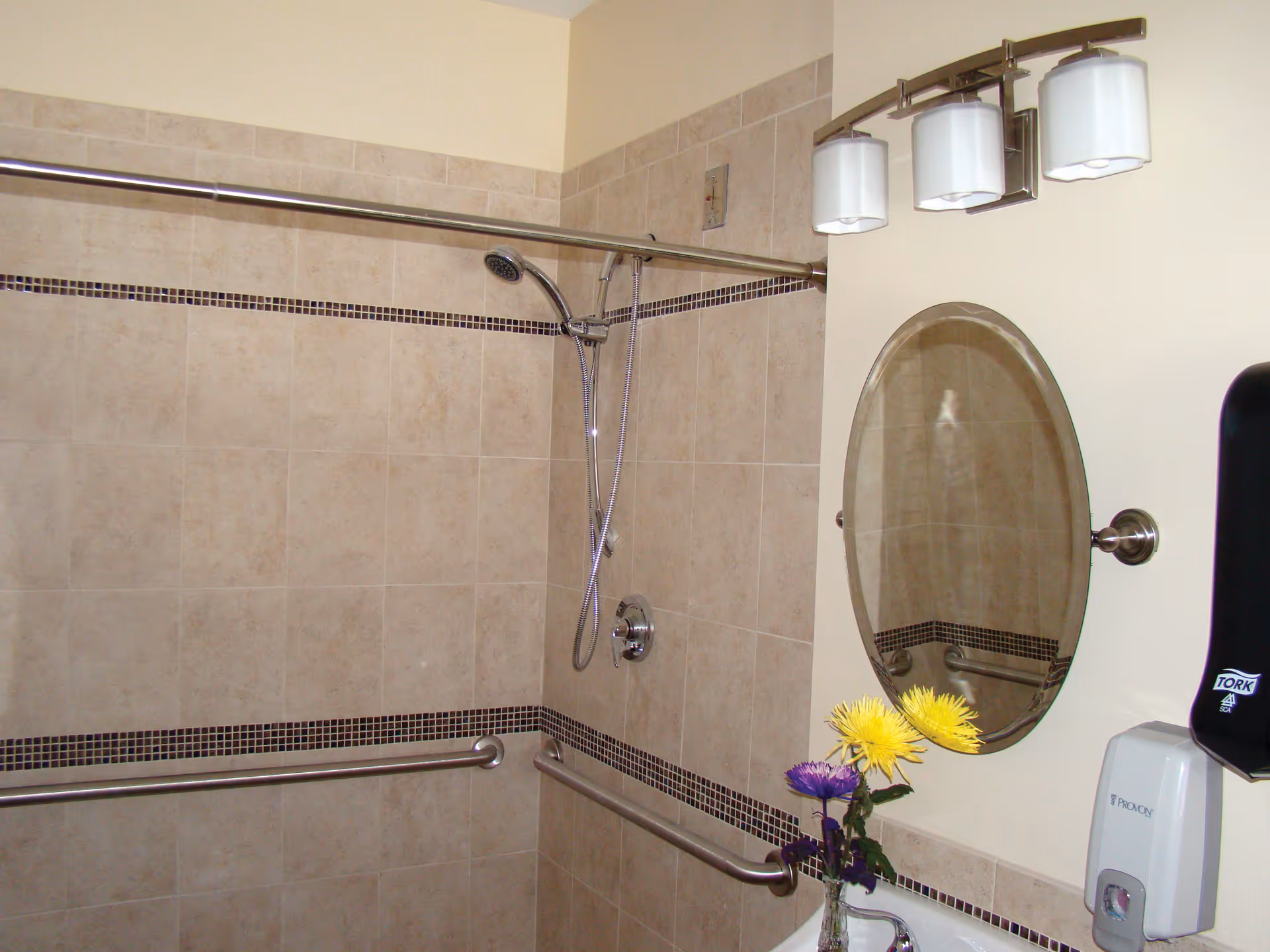A bathroom with beige tiled walls featuring a showerhead and grab bars. There is an oval mirror above a sink with a small vase holding yellow and purple flowers. Above the mirror is a three-light fixture, and to the right is a soap dispenser and a black Tork paper towel dispenser.