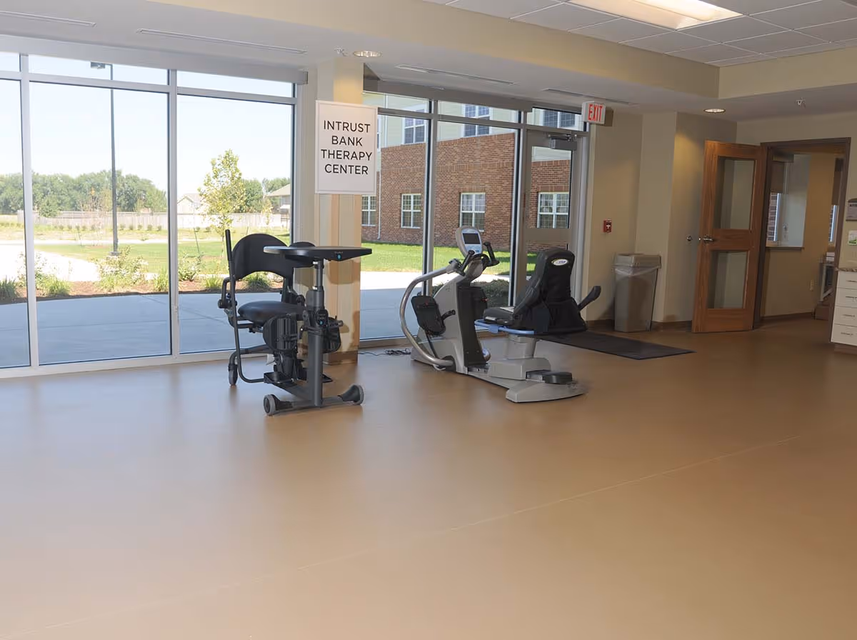 Bright therapy/exercise room with recumbent exercise equipment by large windows and a sign reading 'INTRUST BANK THERAPY CENTER'.