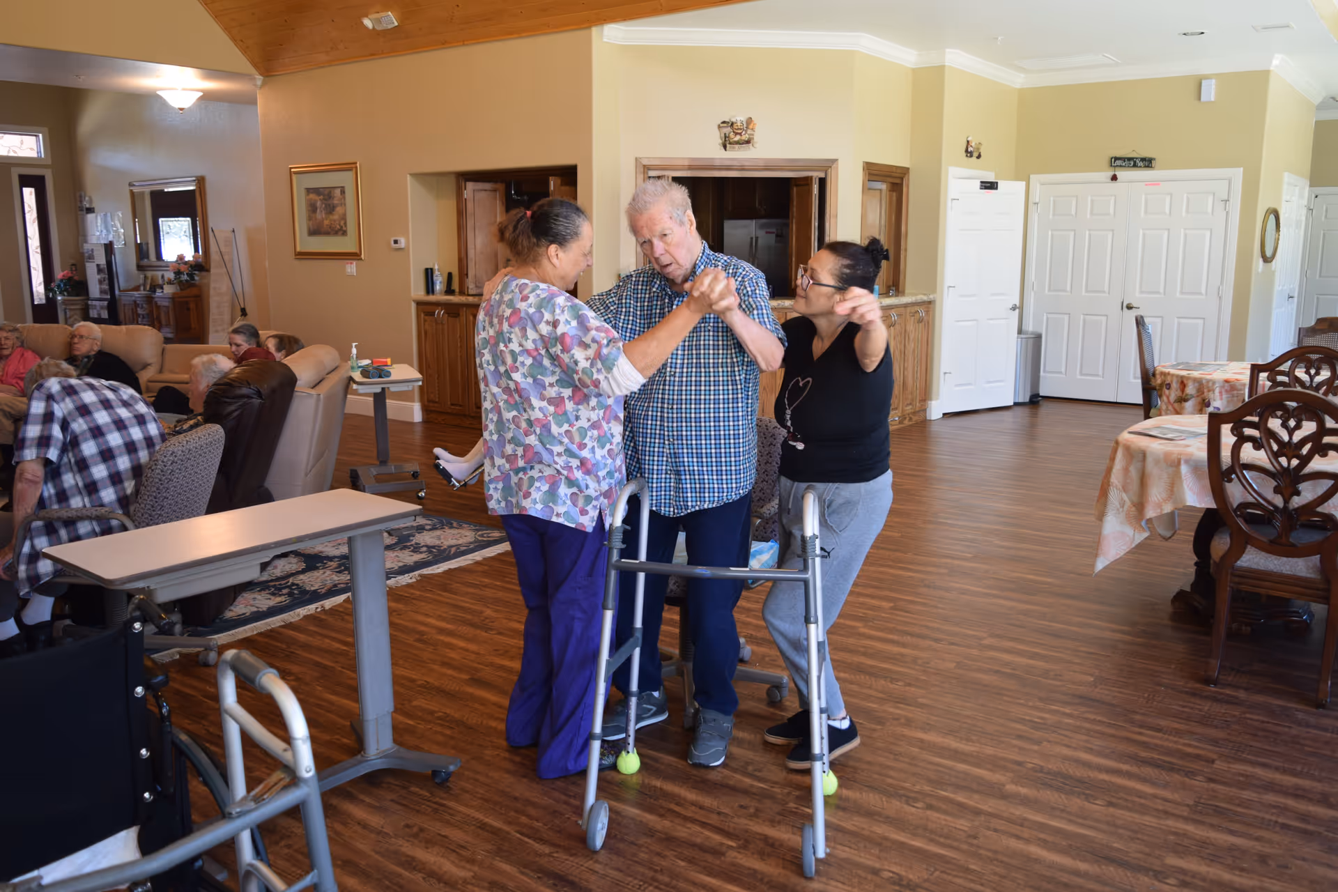 Two women help an elderly man using a walker while other residents sit in a spacious assisted-living common room.