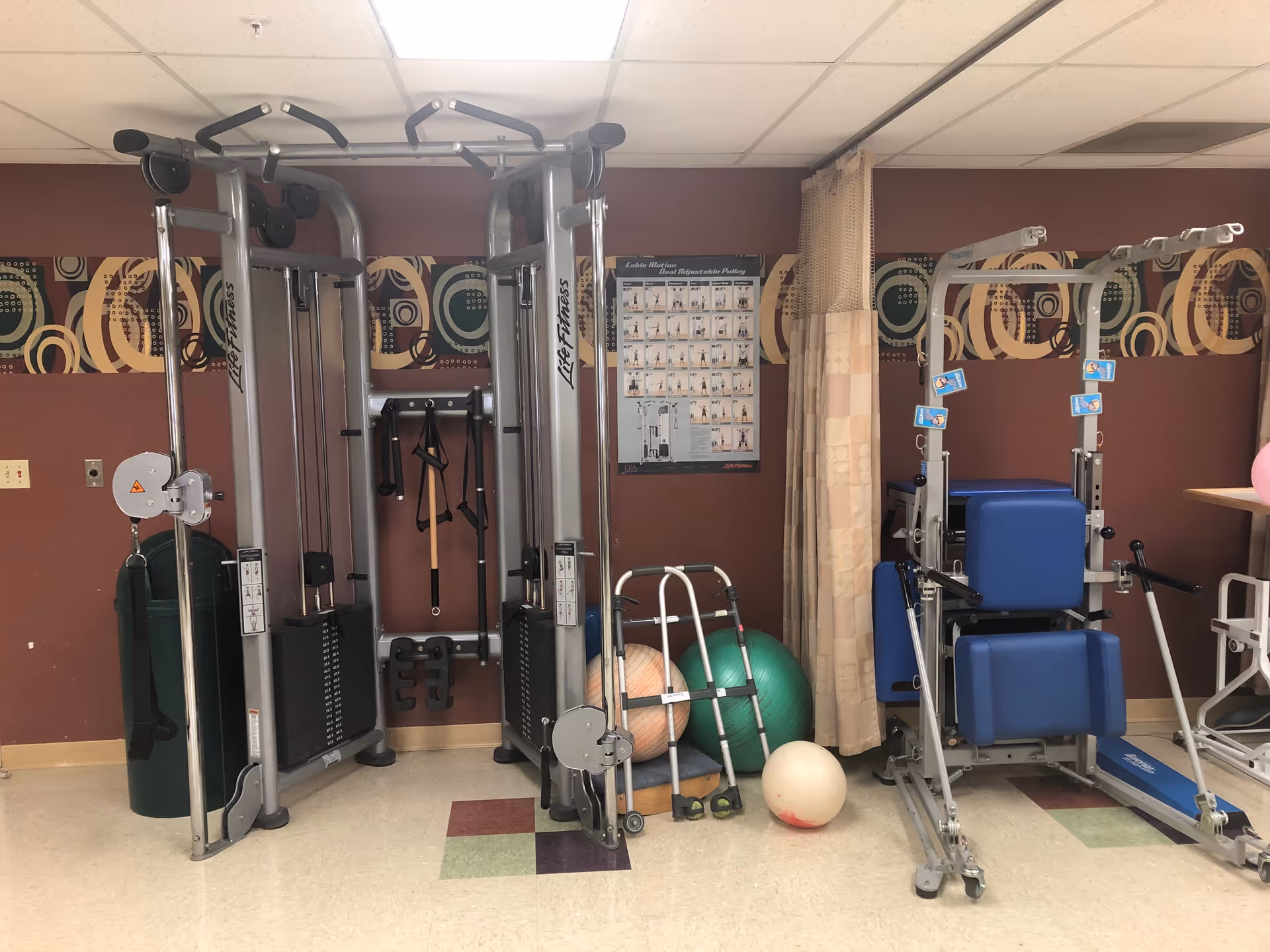 Indoor exercise area with fitness equipment including a Life Fitness dual adjustable pulley machine, a blue padded exercise machine, exercise balls, a walker, and a poster on the wall illustrating cable motion exercises. The room has a patterned wallpaper border and a tiled floor with various colored tiles.