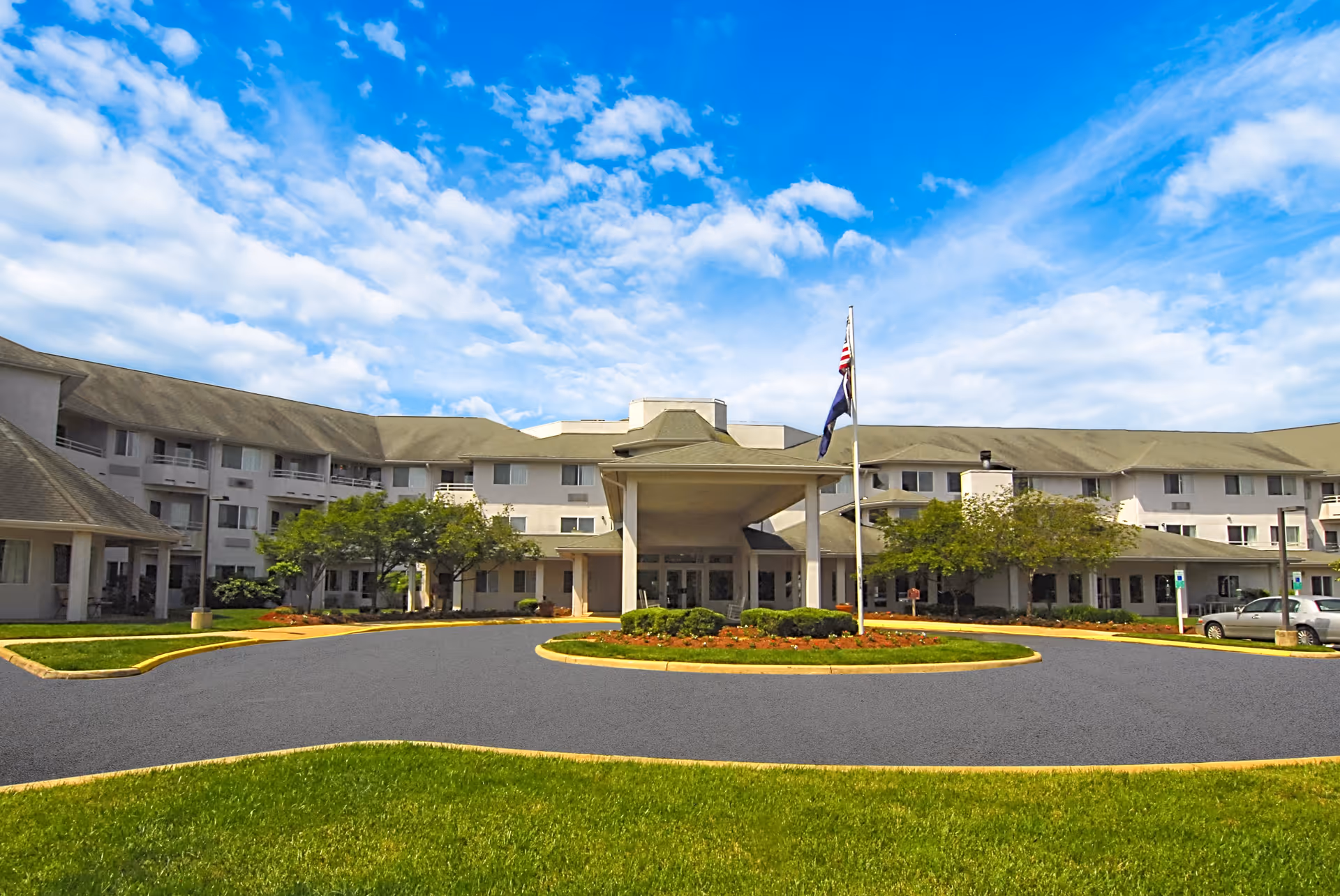 Front exterior view of The Fairmont by Barclay House, a large multi-story senior living facility with a covered entrance, landscaped greenery, and a flagpole with flags under a partly cloudy blue sky.