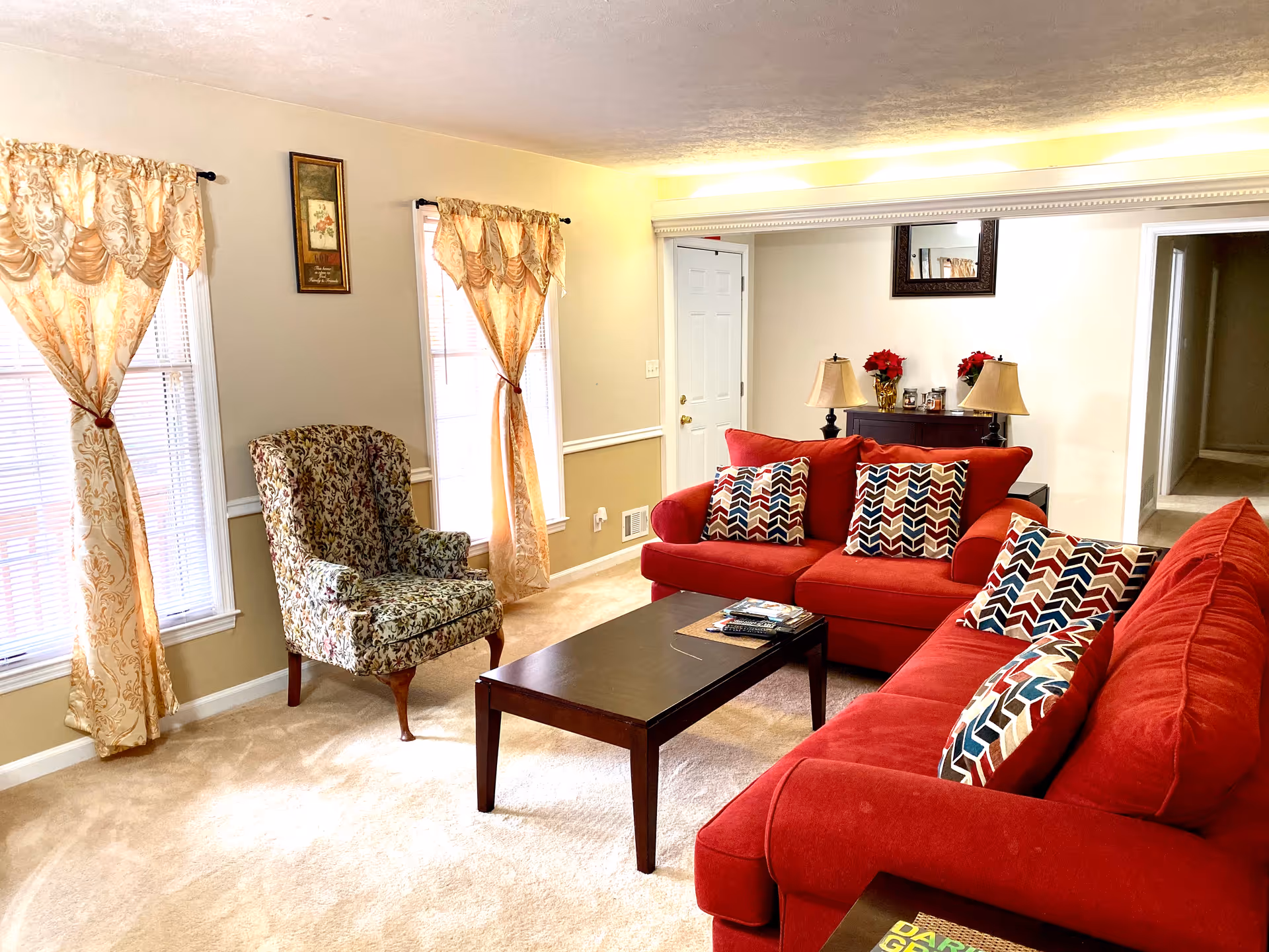 A cozy living room with two red sofas adorned with colorful patterned pillows, a dark wooden coffee table with magazines and remote controls, a floral upholstered armchair near two windows with sheer gold curtains, and a console table with two lamps and red floral decorations beneath a wall mirror.