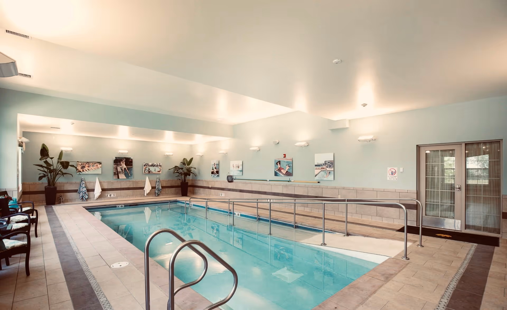 Indoor swimming pool area with handrails and a ramp for accessibility. The pool is surrounded by tiled flooring and light blue walls adorned with framed pictures and potted plants. There are chairs along one side and double glass doors at the far end of the room.