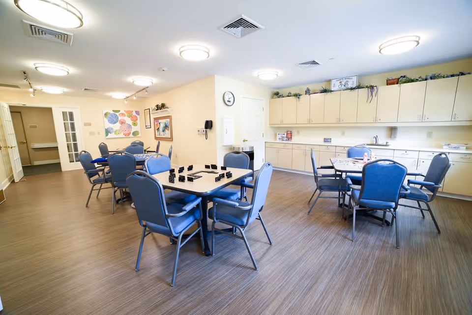 A bright and spacious common room with multiple tables and blue cushioned chairs arranged around them. The room has light-colored walls, a wood-patterned floor, and ceiling lights. There are cabinets along one wall with decorative plants on top, a clock on the wall, and colorful artwork hanging. The tables have board games and other small items on them.