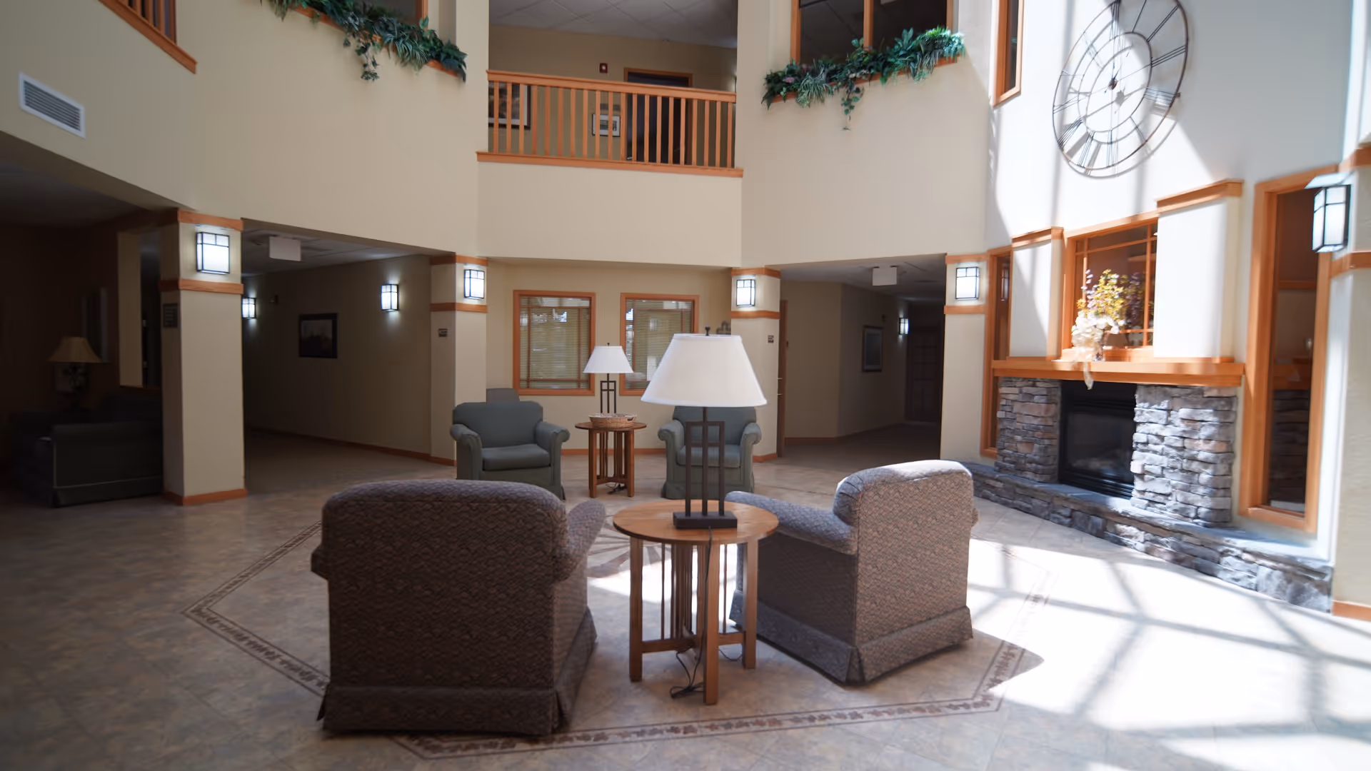 A spacious and well-lit common area in a senior living facility featuring comfortable armchairs arranged around a small wooden table with a lamp. The room has high ceilings with a large decorative clock on the wall above a stone fireplace. There are windows and greenery along the upper balcony railing, and soft natural light fills the space.