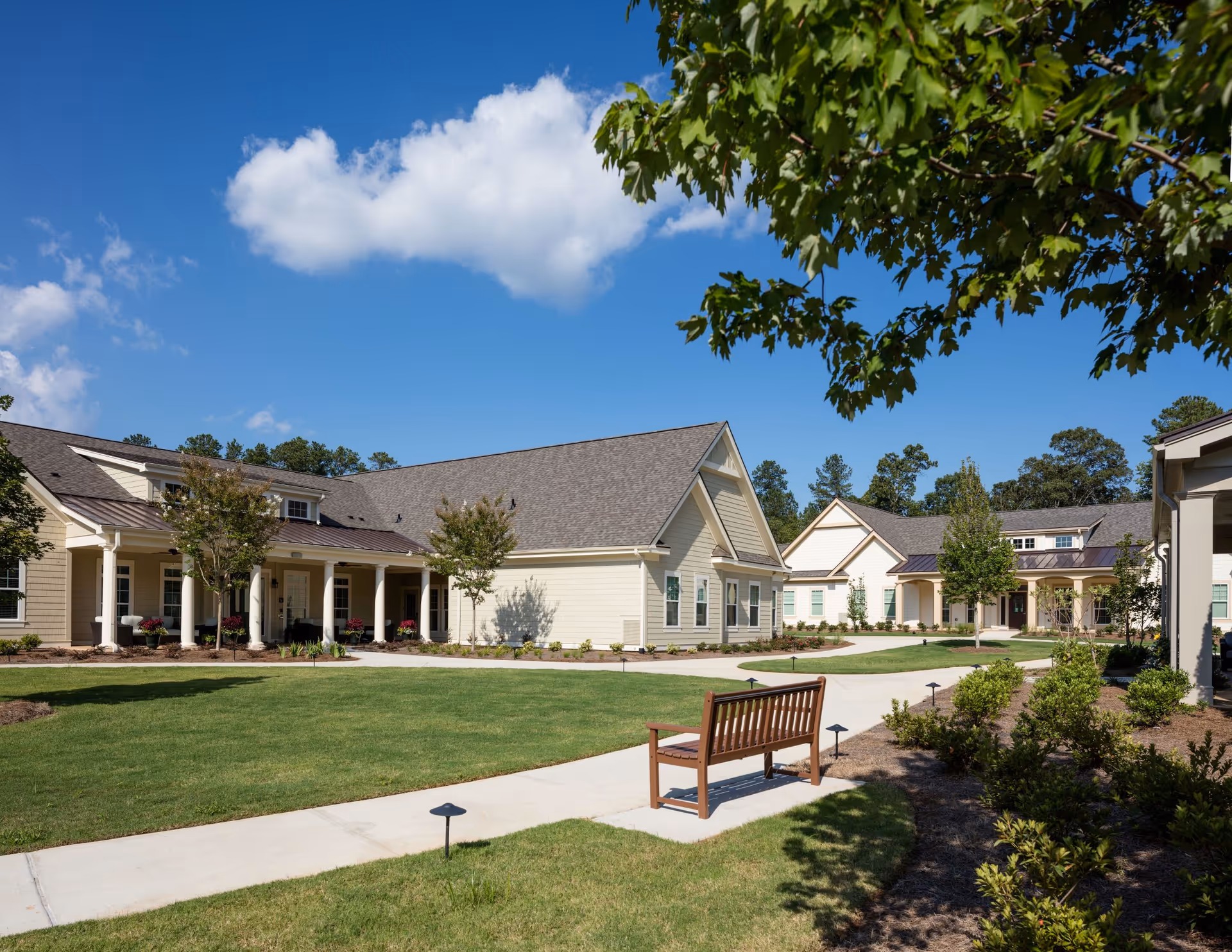Outdoor view of a senior living community with beige buildings featuring pitched roofs and white columns. There is a paved walkway, green lawn, small trees, shrubs, and a wooden bench under a clear blue sky with some clouds.