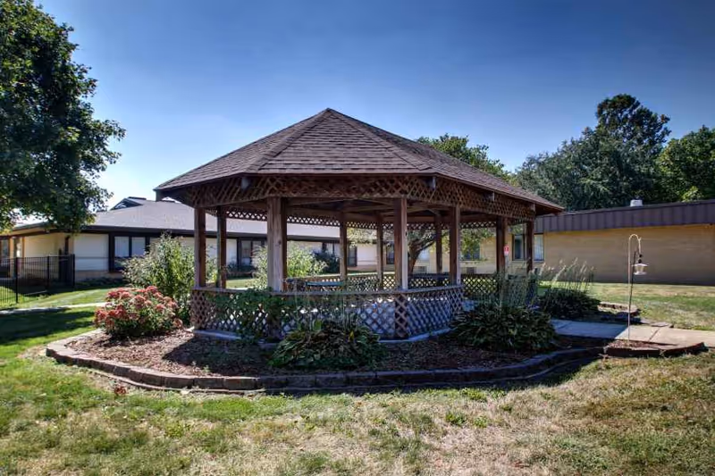 A wooden octagonal gazebo in a landscaped courtyard with grass, shrubs, and surrounding single-story buildings.