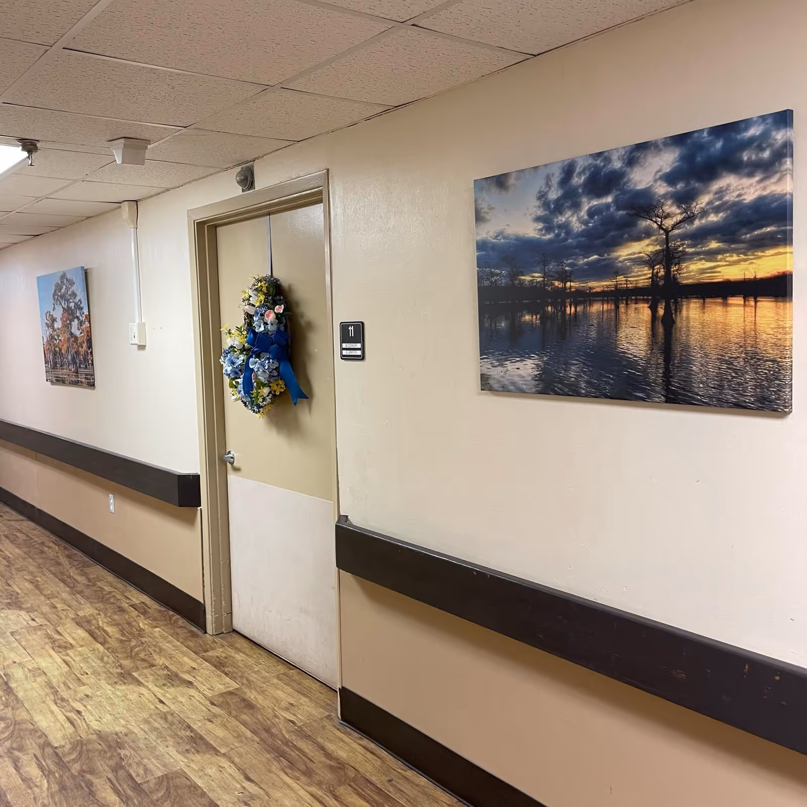 A hallway in a healthcare facility with wood-patterned flooring and beige walls. There is a door labeled with the number 11 decorated with a floral wreath featuring blue and yellow flowers. Two landscape paintings hang on the walls, one showing a sunset over water with trees and the other showing a scene with trees and a fence.