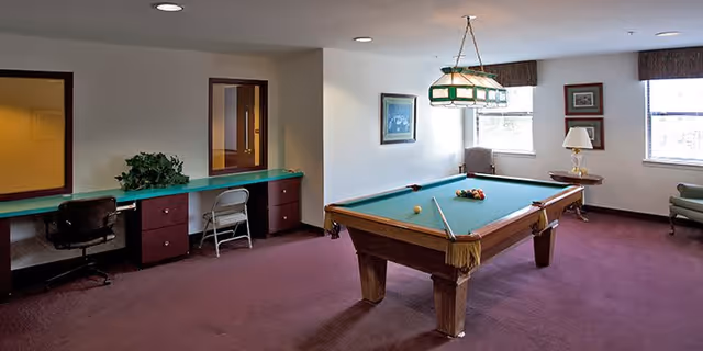 A recreational room with a pool table in the center, a green felt surface, and billiard balls arranged on it. The room has a maroon carpet, two windows letting in natural light, framed pictures on the walls, a green and white hanging light fixture above the pool table, and a long counter with chairs along one wall.
