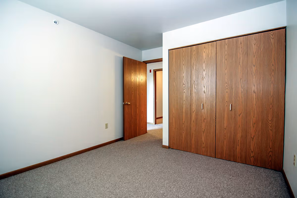 Empty room with beige carpet, white walls, a wooden door partially open, and a large wooden closet with four doors.
