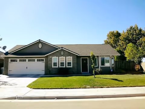 Single-story suburban house with a two-car garage, green lawn, and trees in front under a clear blue sky.