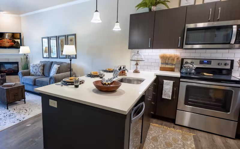 Open-plan kitchen with a central island, stainless steel appliances, and a view into a furnished living area.