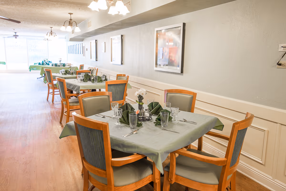 A dining room with several tables covered in green tablecloths and set with glassware, silverware, and folded green napkins. Wooden chairs with green cushions surround the tables. The room has light-colored walls with framed pictures and ceiling lights, and large windows letting in natural light at the far end.