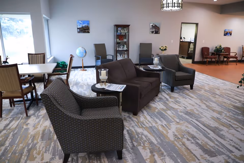 A spacious and well-lit common area in an assisted living facility featuring a brown loveseat and two patterned armchairs arranged around small tables with candles and a newspaper. In the background, there are additional chairs, a globe, a display cabinet with decorative items, and a dining table near large windows letting in natural light.