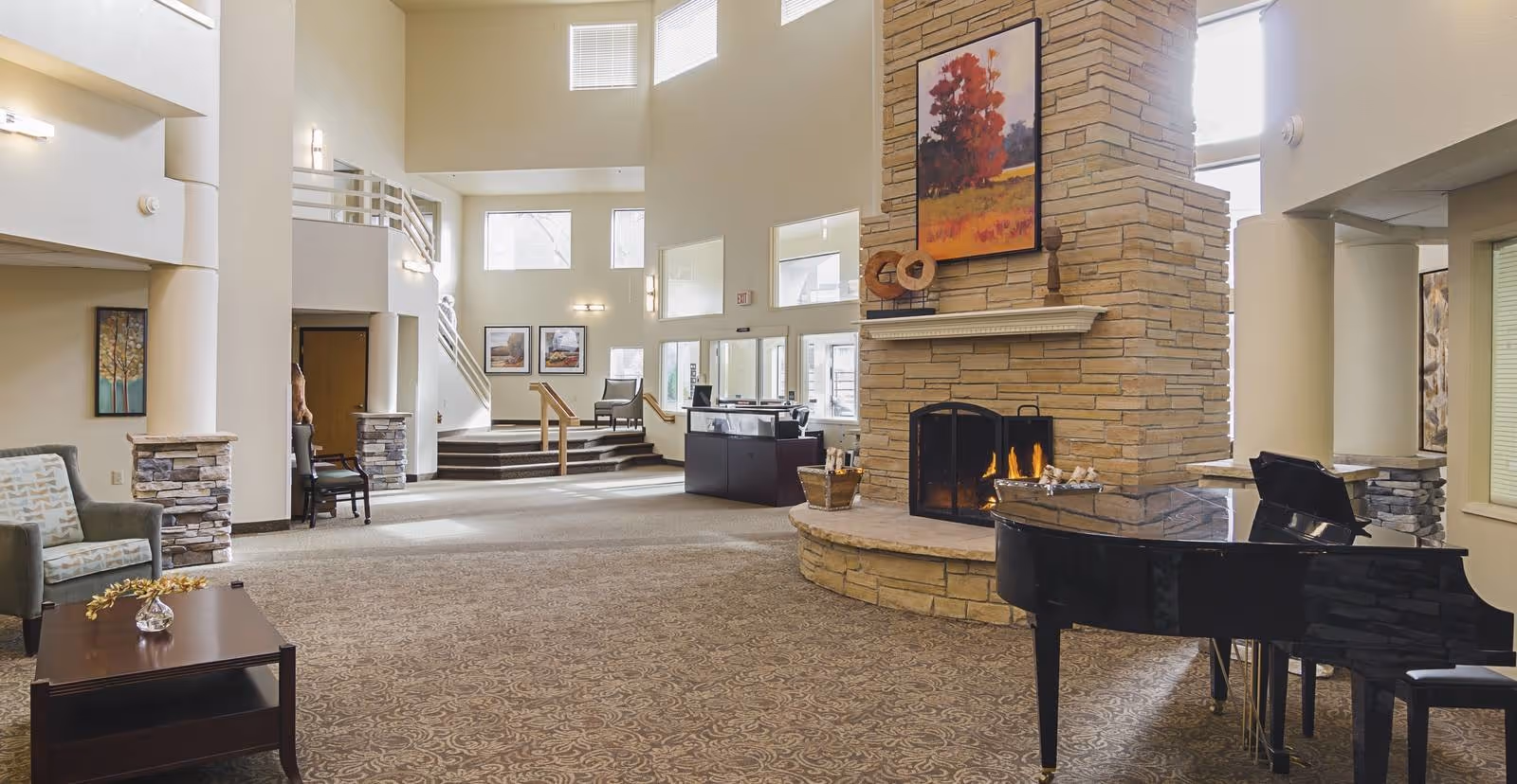 Spacious and well-lit living room area with a large stone fireplace, a black grand piano, comfortable seating including armchairs and a coffee table, and artwork on the walls. The room features high ceilings, multiple windows, and a carpeted floor.