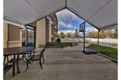 Covered outdoor patio next to a brick building with metal tables and chairs, a portable basketball hoop, raised planters, and a fenced lawn.