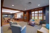 Spacious senior living common room with sofas, armchairs, a coffee table, and a dining area visible in the background.