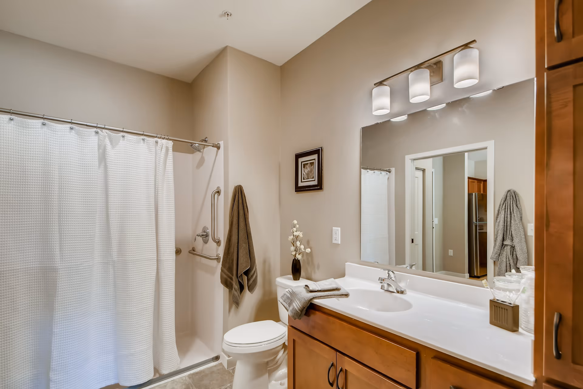 A clean and well-lit bathroom featuring a shower with a white curtain, a toilet with a vase of flowers on top, a wooden vanity with a white countertop and sink, a large mirror above the sink with three light fixtures, and a towel hanging on the wall next to the shower.