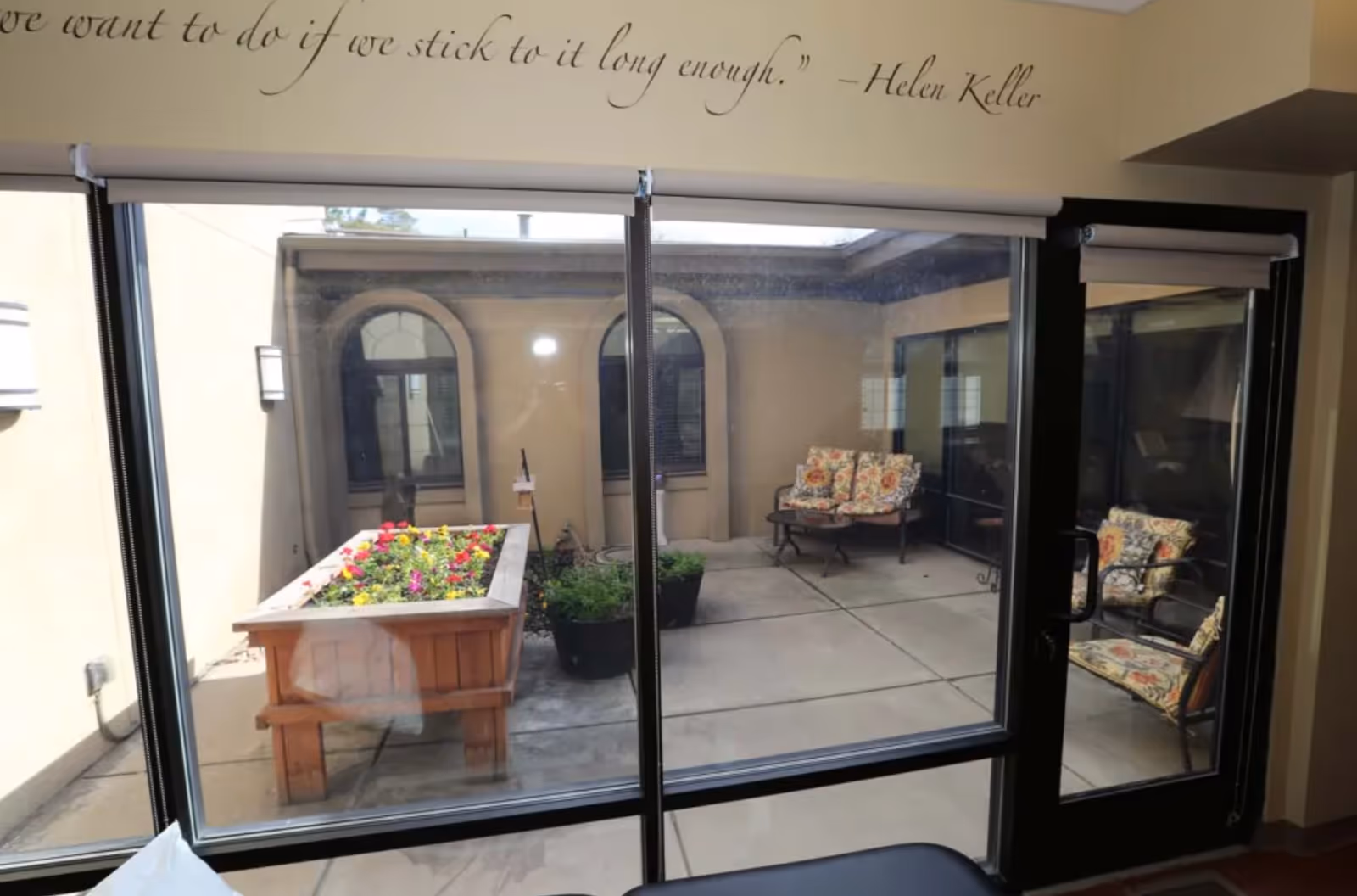 View through large glass windows and a glass door into a small outdoor courtyard with a raised wooden planter box filled with colorful flowers and several potted plants. There are cushioned chairs with floral patterns arranged around the courtyard. The interior wall above the window has a quote by Helen Keller.