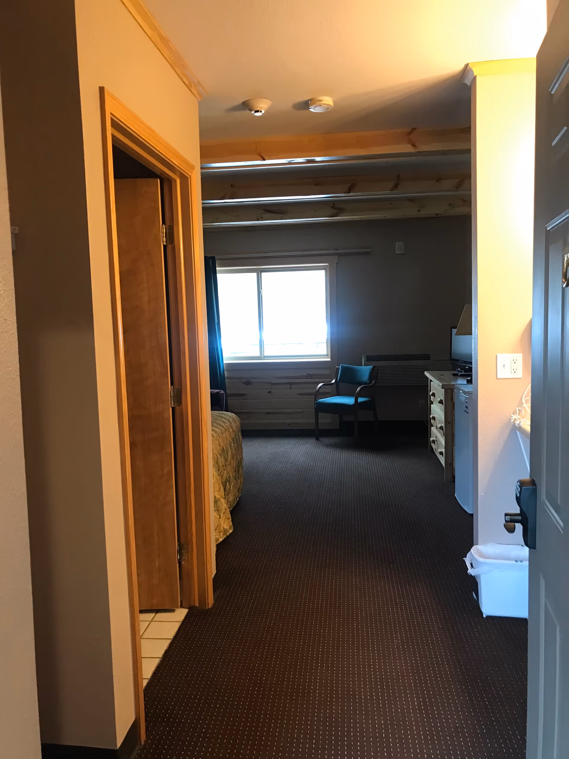 View down a hallway into a room with a window, a chair, a dresser with a small TV, and a bed partially visible. The hallway has a door on the left leading to a bathroom or closet. The room has carpeted flooring and wooden ceiling beams.