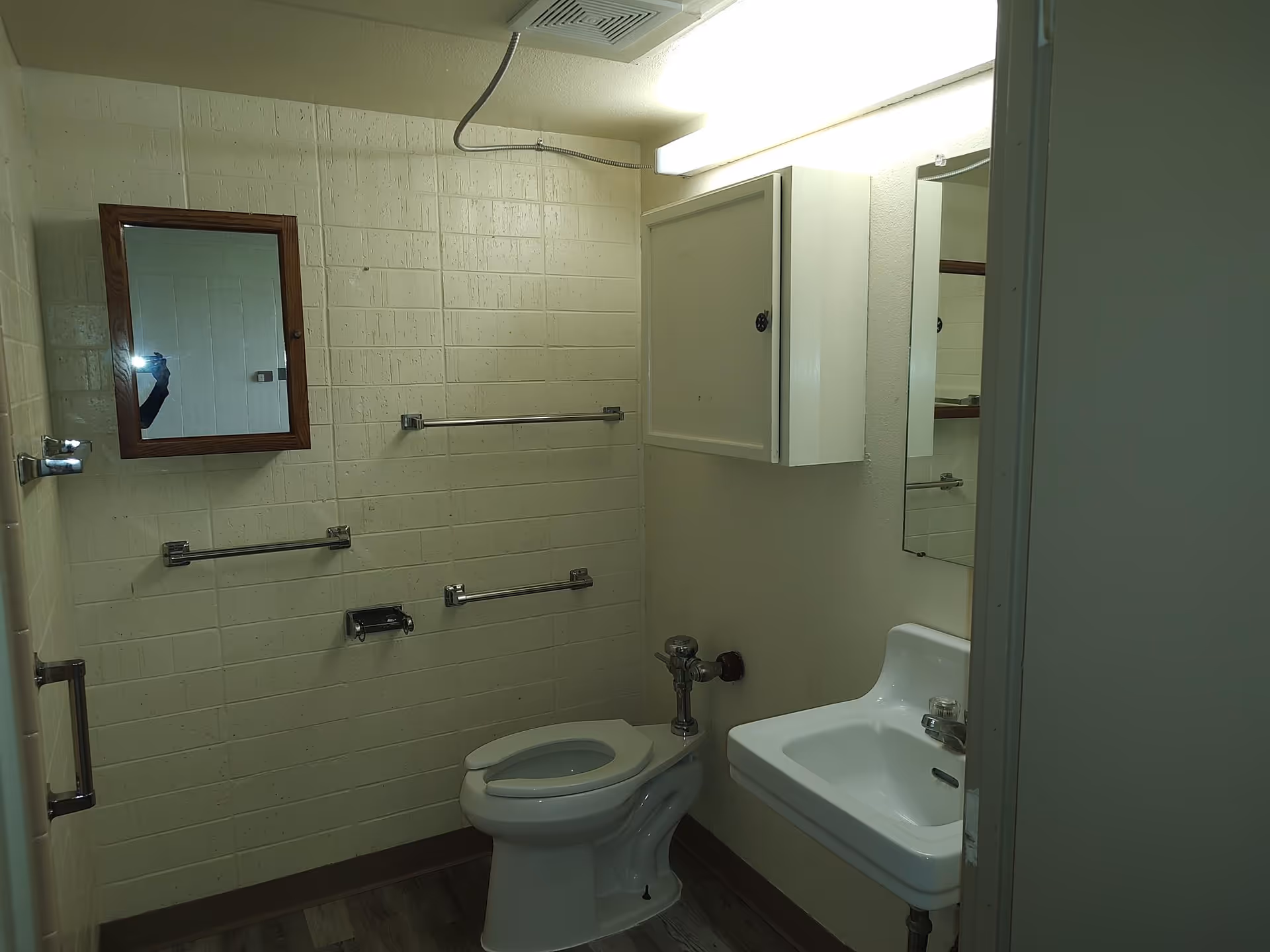 A small bathroom with a toilet, wall-mounted sink, mirrored medicine cabinets, and several towel and grab bars.