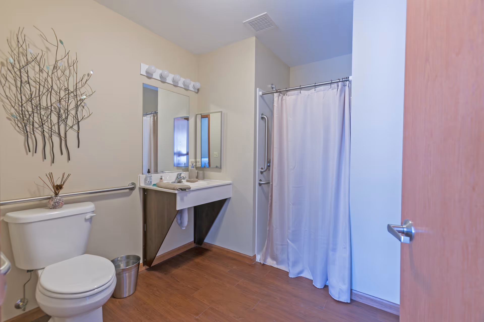 A clean and accessible bathroom featuring a toilet with a metal grab bar, a wall-mounted sink with a mirror above it, a metal trash can, and a shower area with a curtain and grab bars. The walls are light-colored, and there is decorative metal wall art above the toilet. The floor has wood-like flooring.