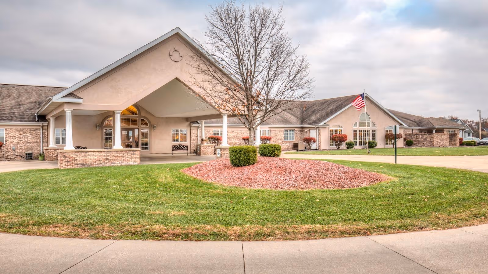 Front exterior view of a single-story senior living facility with a covered entrance supported by white columns, a circular driveway, a landscaped area with grass, mulch, and bushes, and an American flag on a flagpole. The building has beige walls with brick accents and multiple windows.