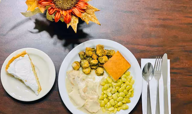 A meal on a wooden table consisting of a plate with sliced turkey in white gravy, fried okra, cornbread, and lima beans. Next to it is a small plate with a slice of pie topped with whipped cream. A set of silverware wrapped in a white napkin is placed to the right of the main plate. A decorative sunflower centerpiece is partially visible at the top of the image.