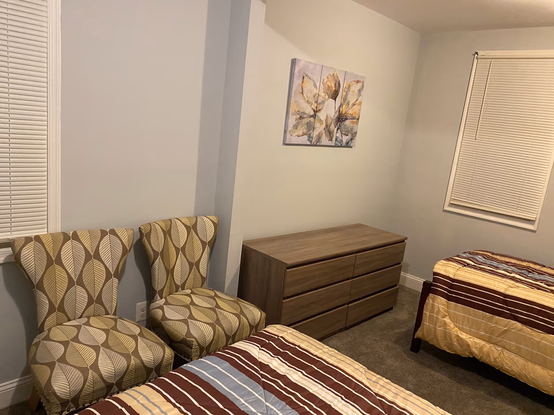 A bedroom with two single beds covered with striped bedspreads in shades of brown, yellow, and blue. There are two patterned chairs with leaf designs placed against the wall near a window with closed blinds. A wooden dresser with six drawers is positioned against the wall, and above it hangs a three-panel artwork featuring abstract leaves. The room has light-colored walls and carpeted flooring.