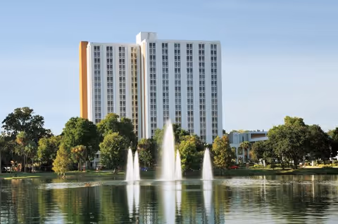 A tall white multi-story building with orange accents behind a pond with three water fountains, surrounded by green trees under a clear blue sky.