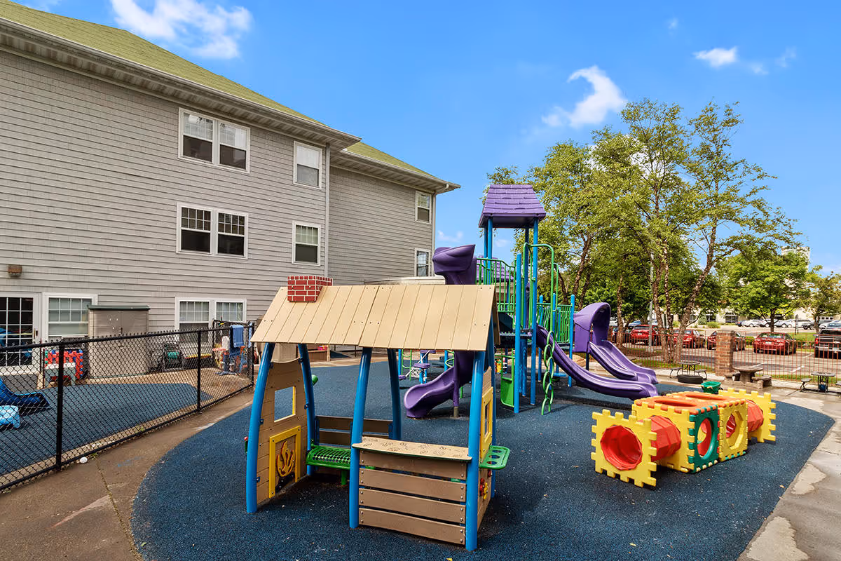 Outdoor playground area with colorful play structures including slides, tunnels, and a small playhouse. The playground is surrounded by a black chain-link fence and is situated next to a multi-story building with gray siding. Trees and parked cars are visible in the background under a blue sky with some clouds.