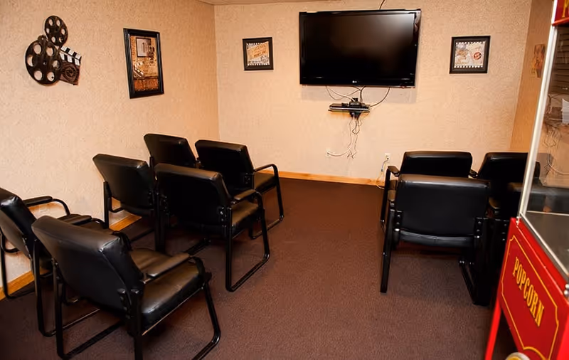 Small room with a wall-mounted flat screen TV, six black cushioned chairs arranged in two rows facing the TV, movie-themed wall decorations, and a red popcorn machine on the right side.