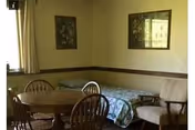 A small room with a single bed covered with a floral bedspread, a round wooden table with four wooden chairs, a cushioned armchair, two framed floral pictures on the wall, and a window with curtains letting in natural light.