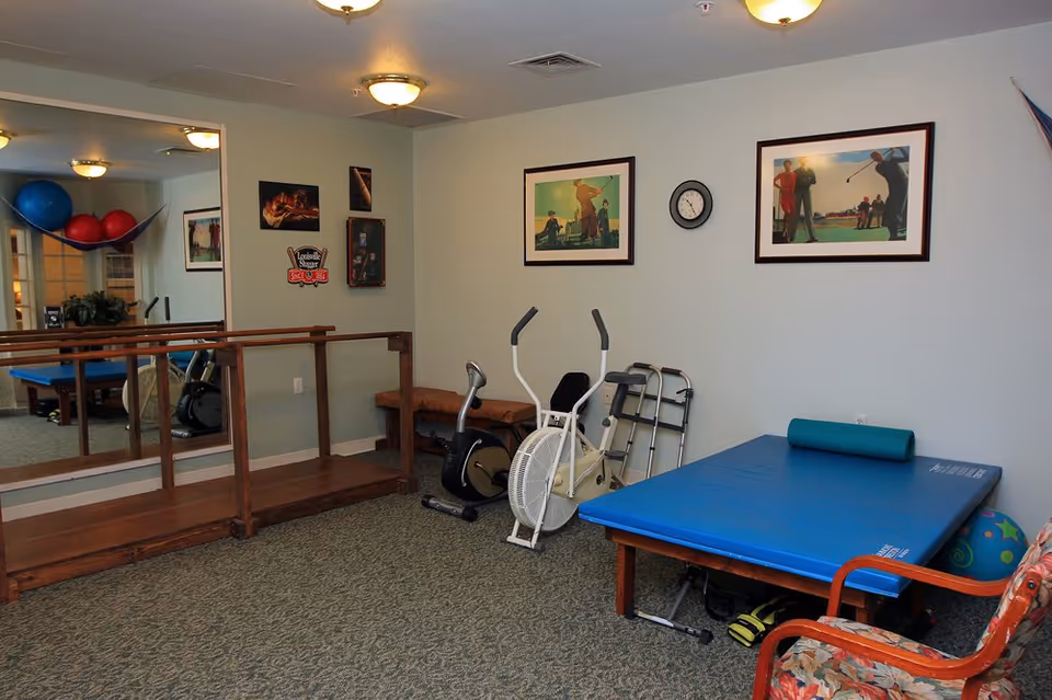A small exercise room with a wooden ramp, exercise bike, walker, blue exercise mat with a foam roller, and a chair with floral upholstery. The walls have framed pictures of people playing golf and a clock. There is a large mirror on one wall reflecting exercise balls and part of the room.