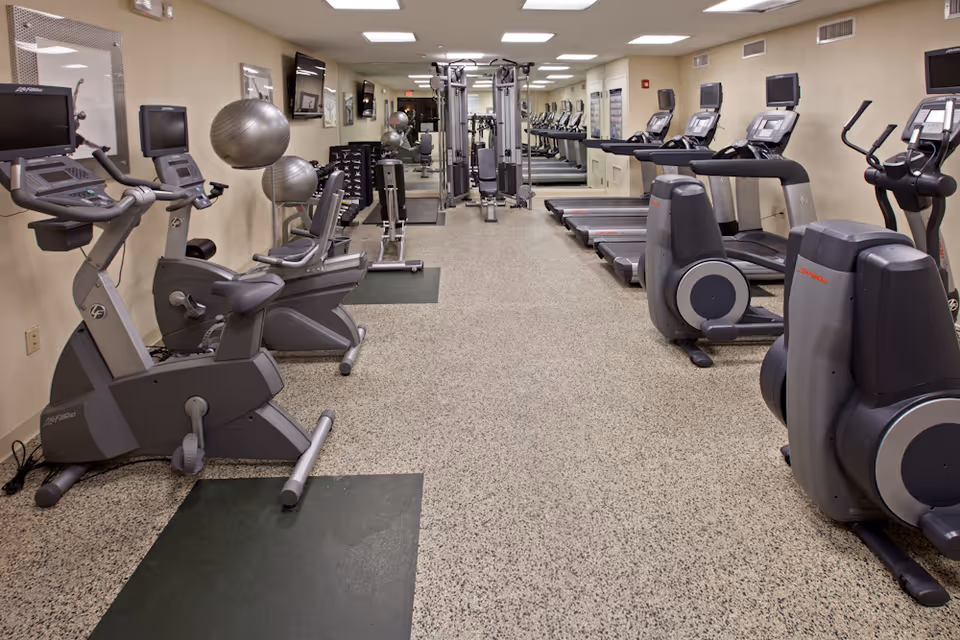 Interior view of a fitness room in an assisted living facility with exercise equipment including stationary bikes, treadmills, elliptical machines, weight machines, and exercise balls. The room has a speckled floor, beige walls, and mirrors on one side.