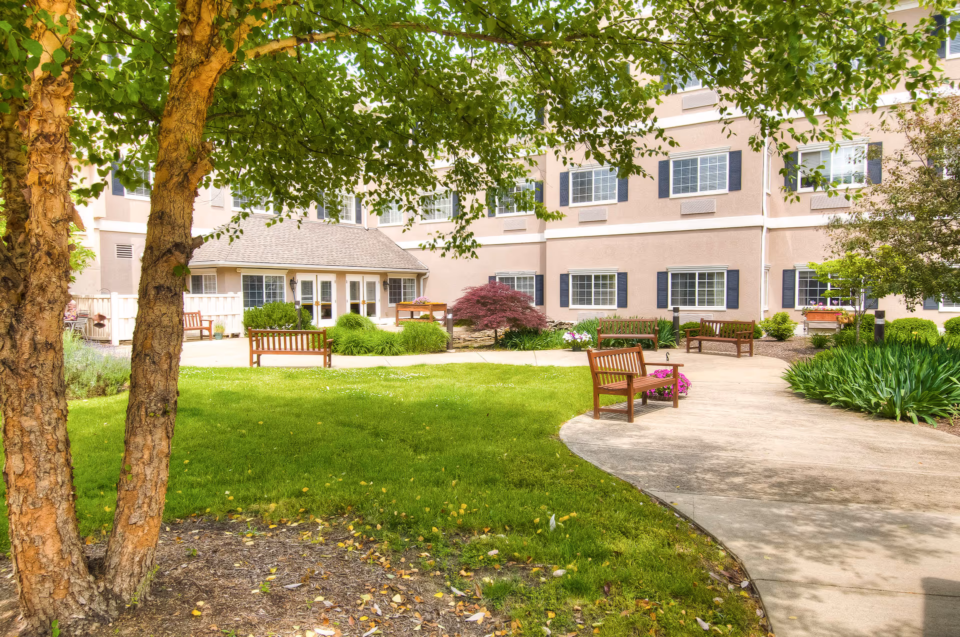 Outdoor courtyard area at The Abbewood featuring green grass, a curved concrete walkway, several wooden benches, various plants and trees, and a multi-story building with multiple windows in the background.