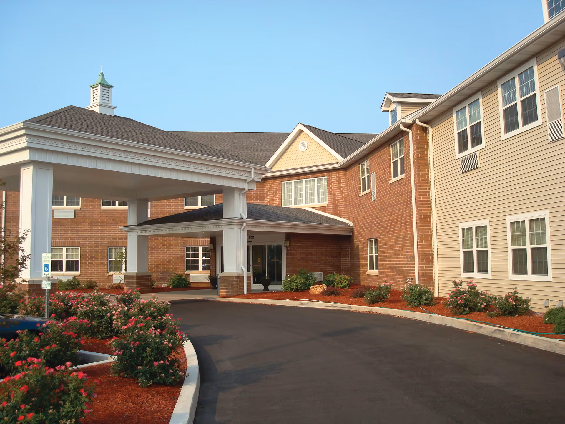 Exterior view of a senior living facility with a covered entrance, brick and siding walls, multiple windows, and landscaped flower beds with red mulch and blooming bushes along a curved driveway.